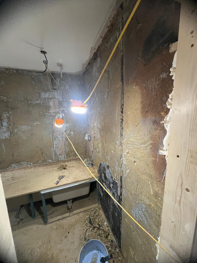 A bathroom during the initial renovation phase with the old suite removed, showing exposed floorboards, bare brickwork, and a timber-framed partition wall.