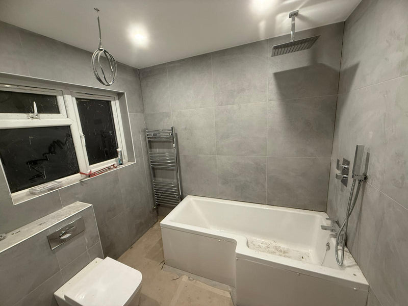 A full bathroom renovation in progress, featuring large-format grey wall tiles, an L-shaped shower bath, wall-hung toilet, and modern chrome fixtures.