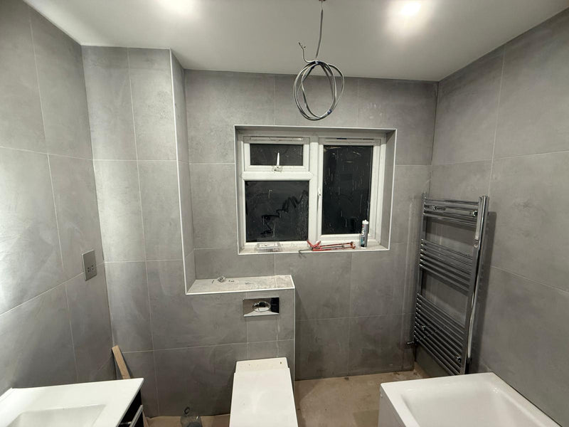 Finished bathroom detail showing a chrome flush plate for a concealed cistern, a modern white toilet, and precision-mitered grey wall tiles around a window reveal.