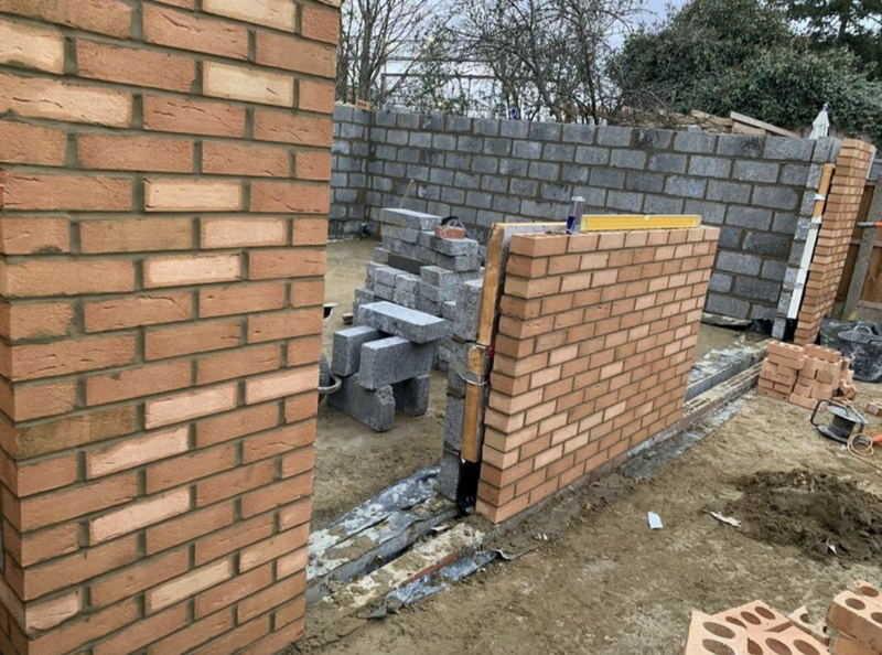 A structural brickwork project in progress, showing a double-skin wall with external red facing bricks and internal grey breeze blocks. A yellow spirit level is used to ensure perfect vertical alignment, demonstrating expert masonry for a London residential extension.