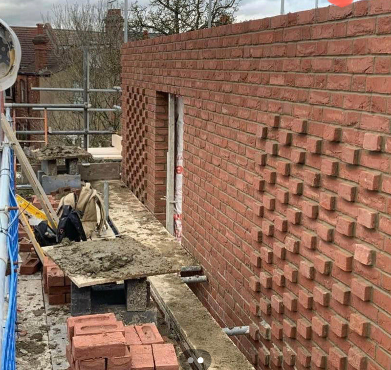 A cross-section of a house extension wall under construction. The image shows the external red brick skin, the internal structural breeze blocks, and the high-quality cavity wall insulation being installed to meet London building regulations.