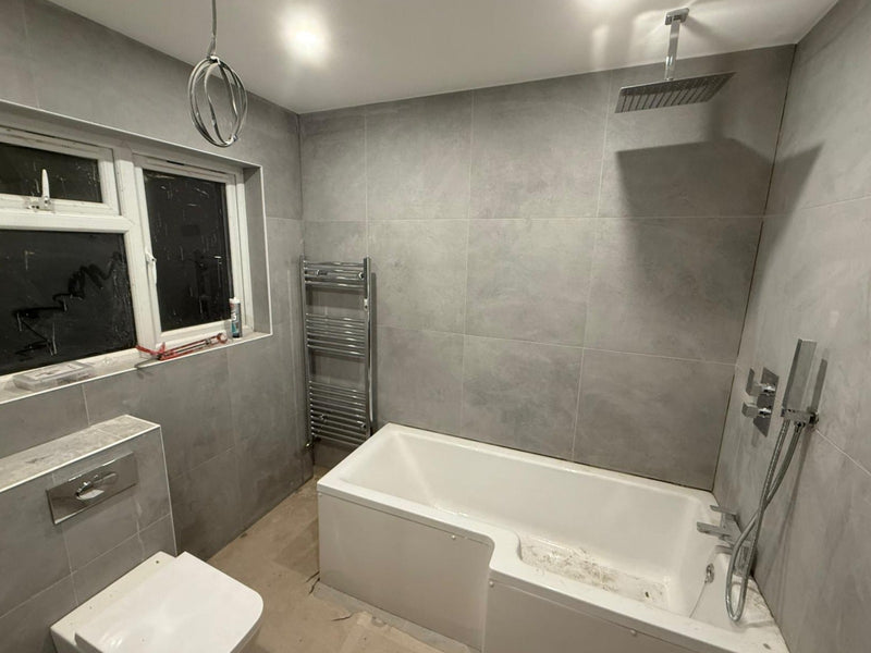A completed modern bathroom featuring an L-shaped shower bath with a rainfall head, grey concrete-effect wall tiles, and a wall-hung toilet.