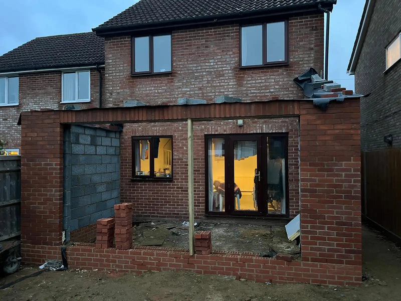 A rear house extension under construction in London, showing the red brickwork shell, structural lintels, and breeze block walls. The image highlights the professional site layout and the transition between the existing property and the new expansion.