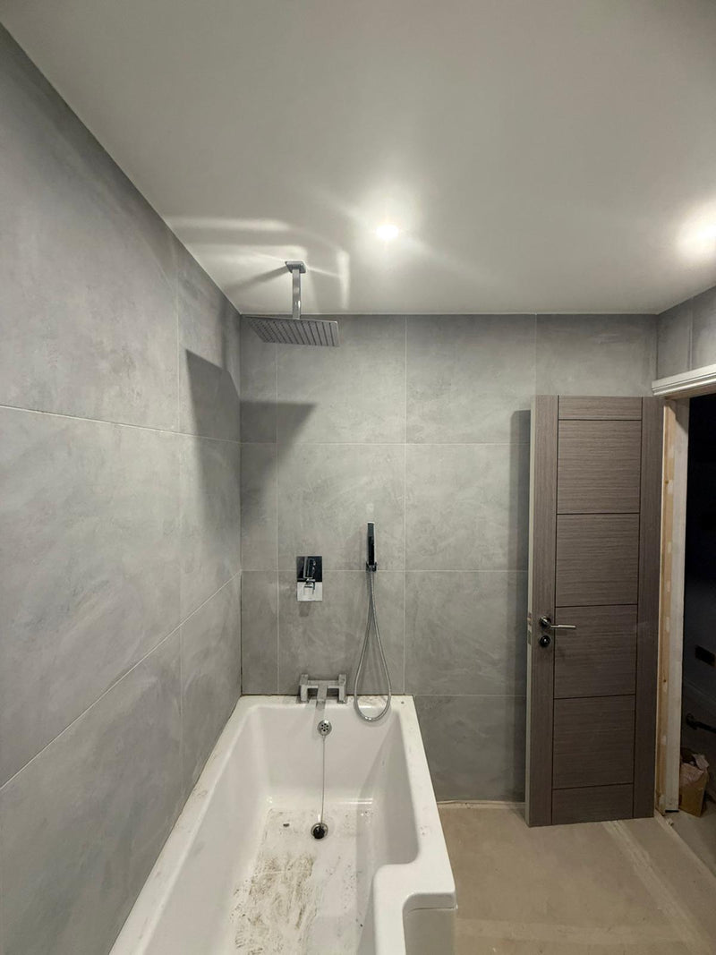 A modern bathroom featuring a white shower-bath combo, a ceiling-mounted rainfall shower head, grey large-format wall tiles, and a contemporary grey internal door.