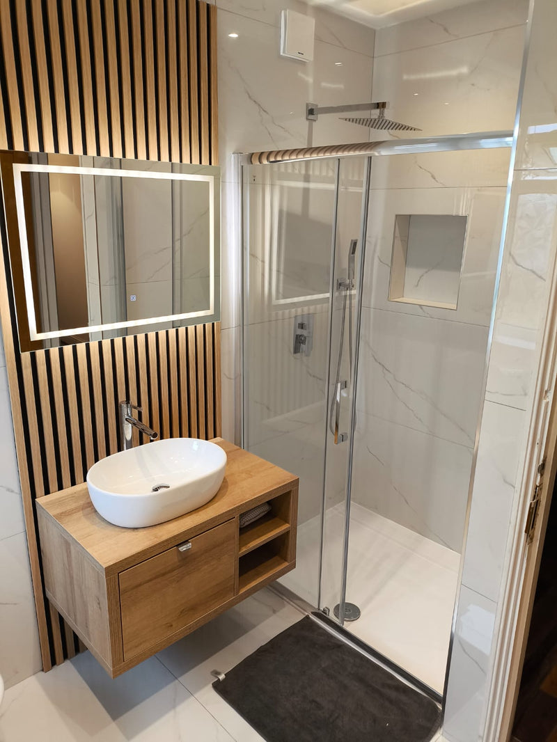 A contemporary bathroom featuring a wood-slat feature wall, a white vessel sink on a floating oak vanity, an illuminated mirror, and marble-effect porcelain shower tiling.