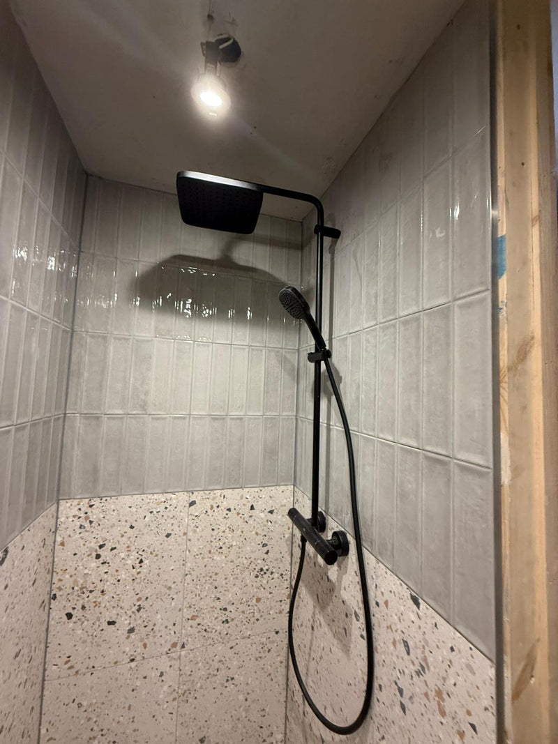 A modern shower featuring a matte black dual-head rainfall shower system, vertical stacked grey metro tiles, and large-format terrazzo base tiling.