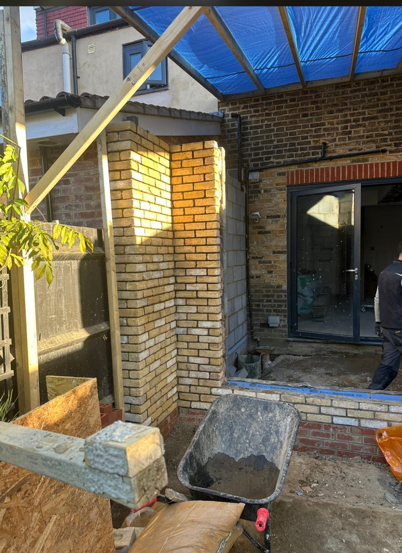 A construction site for a rear house extension featuring a newly built yellow multi-stock brick pillar. The masonry is being integrated with an existing brick elevation and features a modern dark-framed bifold door opening. A temporary timber-framed roof with blue tarpaulin protects the site, which includes a clean mortar-filled wheelbarrow and staged building materials in the foreground.