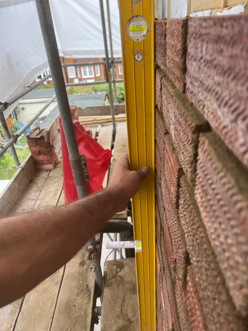 A vertical spirit level held against a newly laid red brick wall on scaffolding, demonstrating perfectly plumb masonry construction.