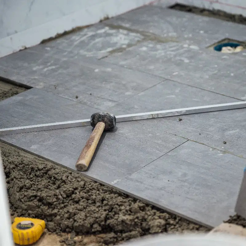 A close-up of grey porcelain floor tiles being installed on a mortar bed. A rubber mallet and a metal spirit level rest on the tiles, showing the precision and leveling process used during a professional bathroom tiling project.