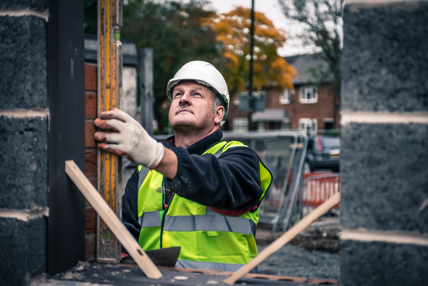 A professional construction worker wearing a white hard hat and high-visibility safety vest using a yellow spirit level to check the vertical alignment of a brick wall on a London residential building site.