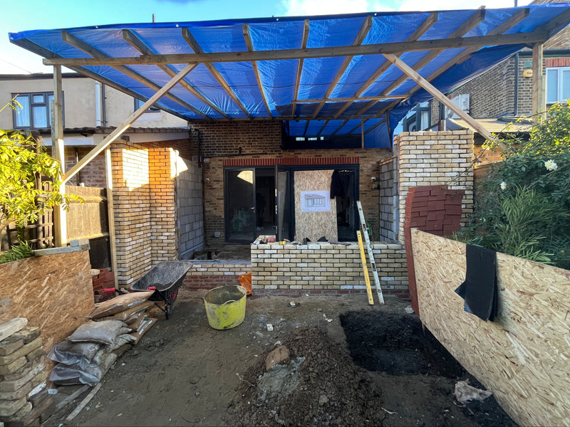 A professional construction site showing a rear house extension under a temporary timber-framed roof with blue waterproof tarpaulin. The image showcases new yellow multi-stock brickwork and internal blockwork pillars being built in a clean, organized workspace with site-safety boarding and staged materials.