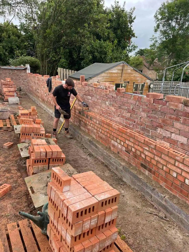 A skilled London bricklayer building a long, red brick garden boundary wall. The image shows a large-scale masonry project in progress with stacks of bricks on pallets, a concrete foundation, and the builder using a trowel to ensure precise brick placement.