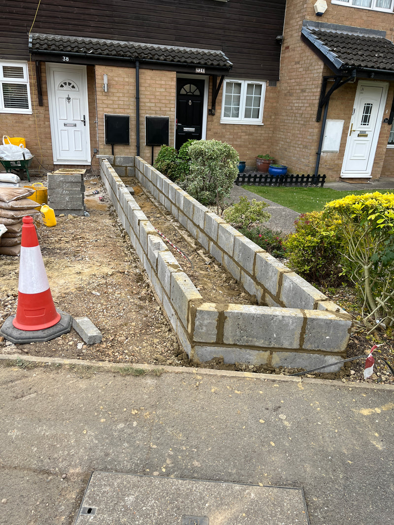 Site view of a residential front garden under renovation, showing the construction of a double-skin hollow block retaining wall for a new planter or boundary.