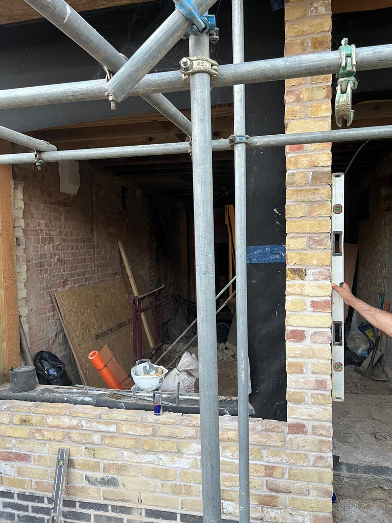 A London home renovation site featuring new brickwork being constructed alongside existing structural walls. The image shows professional scaffolding and a mason using a spirit level to ensure the vertical accuracy of a new brick pillar for a residential extension.