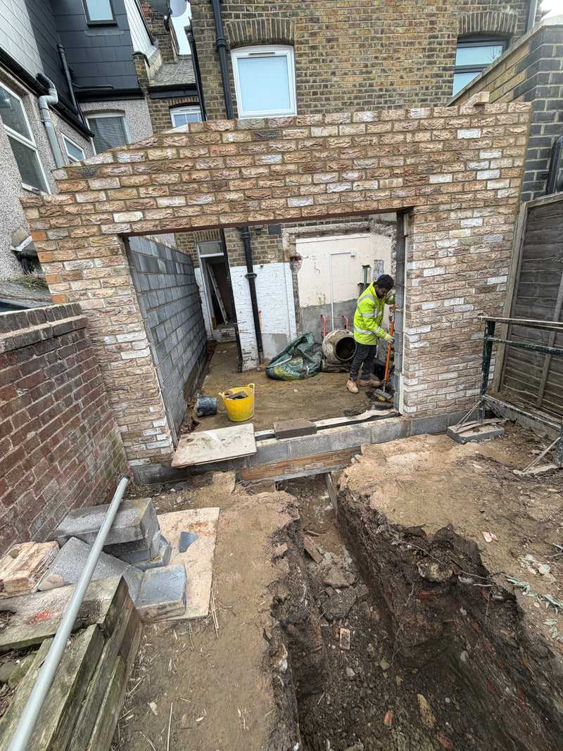 A residential extension under construction in London, showing deep foundation trenches, internal structural blockwork, and a finished brick outer skin.