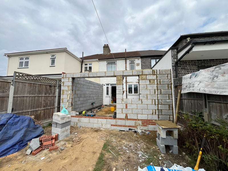A wide-angle view of a rear residential extension during the structural shell phase. The image shows the internal blockwork leaf with large precision-cut openings for a bifold door and window, supported by a red brick engineering plinth. The clean site shows a mix of existing white-rendered property and the new masonry addition.